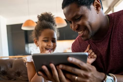 A father and his young daughter smiling at a tablet together