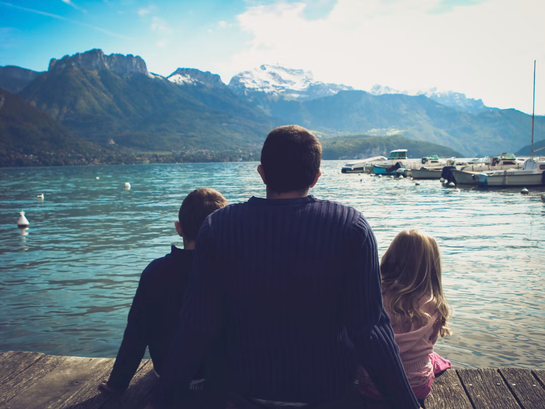 A father and his two children sitting on a jetty looking out over a mountain lake